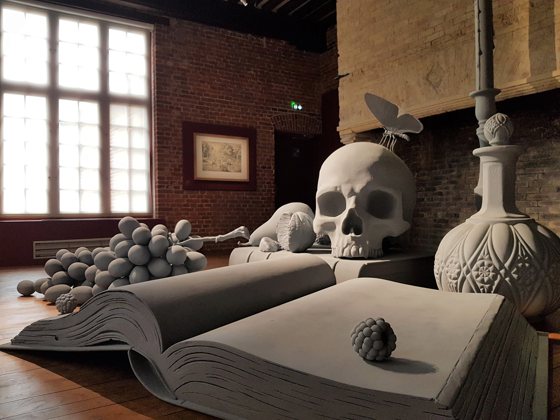 Hans Op de Beeck, Vanitas XL, vanité posé sur le sol du musée avec livre ouvert, bougeoir, crâne, fruits, papillon, unifiés par la couleur grise, exposition Silence & Résonance, Musée de Flandre, Cassel, France, 2023, Boombartstic Art Magazine, Bruxelles