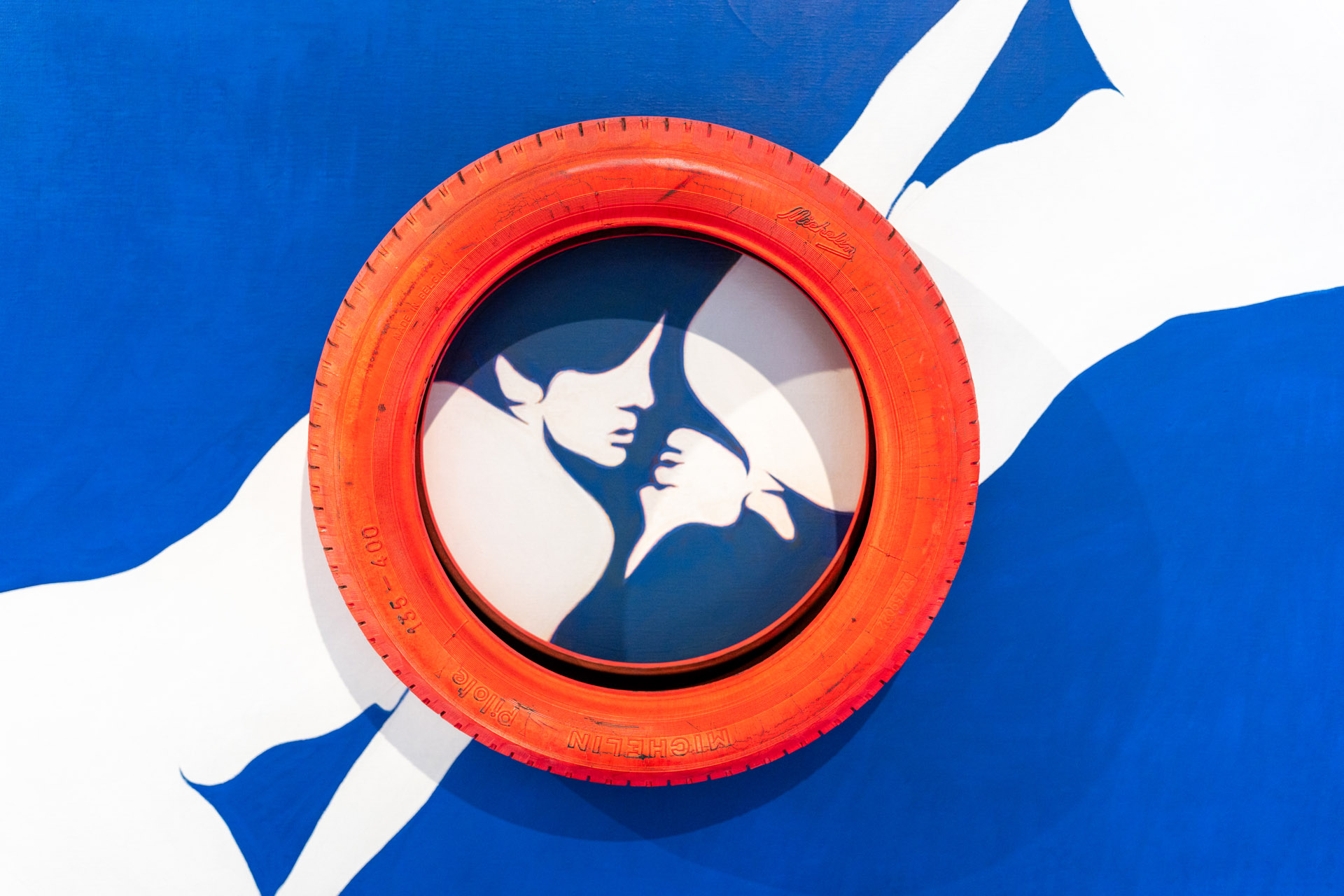 Evelyne Axell, Erotomobile, 1966, deux femmes pop art blanche en tête bêche sur fond peint en bleu. Les visages échangent presque un baiser et son encerclé par un pneu en caoutchouc peint en orange fixé sur la toile, exposition En Regard, Le Delta, Namur, 2023, Boombartstic Art Magazine